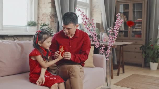 赤いシャツを着た幸せなアジアの父親の肖像画はソファに座って 伝統的なドレスで彼の5歳の娘に中国の旧正月のポストカードを与えます 彼らはカメラに向かって笑っている — ストック動画