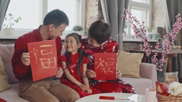 ソファに座って手作りの旧正月のはがきをカメラに見せ 伝統的な服を着てかわいい5歳の女の子と幸せな中国の家族の肖像写真 — ストック動画