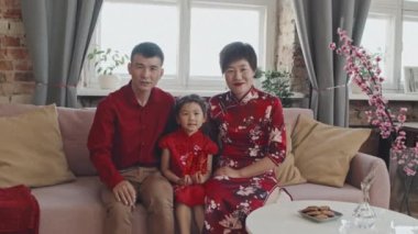 Asyalı ebeveynlerin portresi ve geleneksel kostümler giyen 5 yaşındaki bir kız evdeki koltukta oturup Çin Yeni Yıl kutlamalarında kameraya poz veriyor.
