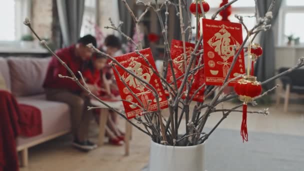 追踪调查镜头集中在明信片和挂在花瓶枝上的中国新年装饰品上 背景为儿童制作明信片的难以辨认的亚洲家庭 — 图库视频影像