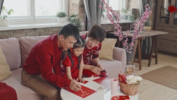 中国の旧正月はがきを作る伝統的なドレスで彼らのかわいい5歳の娘を助けるアジアの両親の中のショット — ストック動画