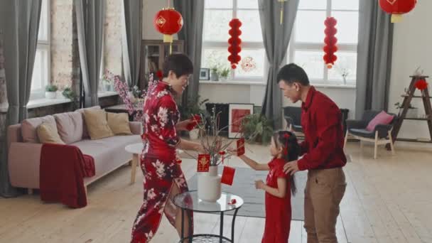 5歳の娘が伝統的な服を着て中国の旧正月の飾りを花瓶に枝に飾り 提灯で飾られたリビングルームで幸せなアジアの家族の中のショット — ストック動画