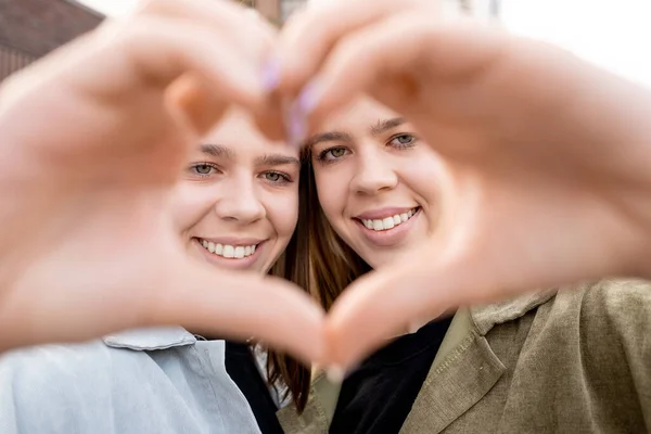 Caras de duas irmãs gêmeas felizes olhando para você através do coração da mão — Fotografia de Stock