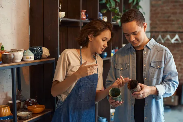 Junge Handwerkerin zeigt männlicher Kundin zwei handgefertigte Becher — Stockfoto