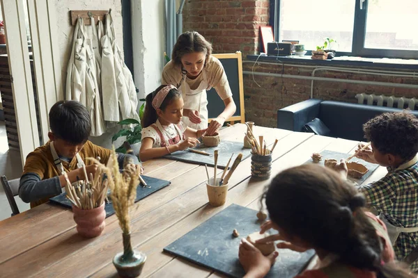 Young craftswoman helping one of schoolkids with making earthenware — Stock Photo, Image