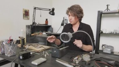 Orta boy kadın kuyumcunun tezgahta oturup büyüteçle bakarken metal yüzüğe lehim yaparken görüntüsü.