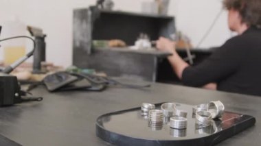Masanın üzerinde duran metal halkaların PAN yakın çekim görüntüsü. Arka plandaki tezgahta, tanınmayan kadın kuyumcu testeresini kullanıyor.