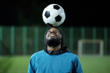 Spor üniformalı sakallı siyah adam alnındaki futbol topuna bakıyor.