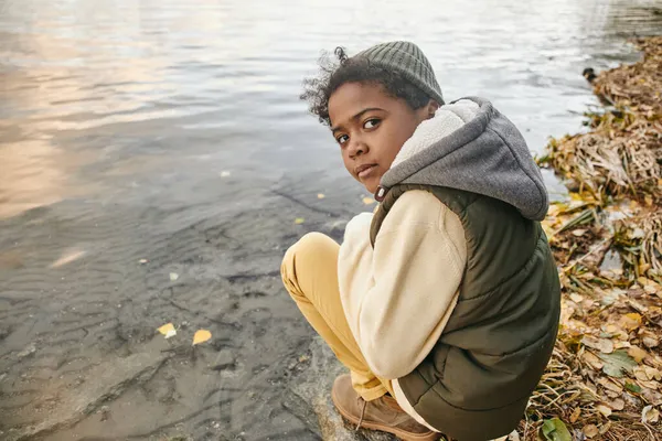 Netter junger Junge in warmer Freizeitkleidung sitzt auf Kniebeugen am Wasser — Stockfoto