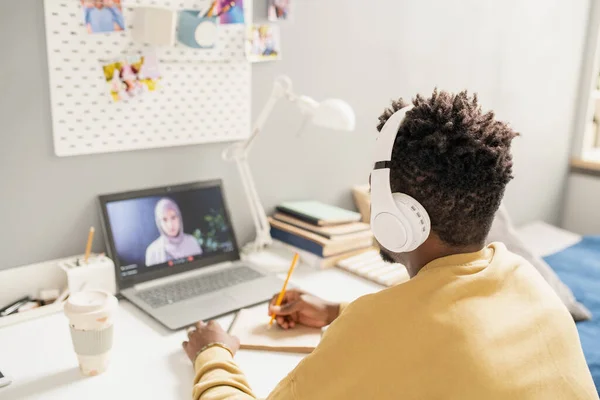 Joven estudiante africano en auriculares sentado delante de la computadora portátil con el profesor en la pantalla —  Fotos de Stock