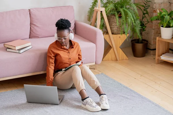 Jovem mulher negra grave em casualwear e fones de ouvido sentado no chão na frente do laptop — Fotografia de Stock
