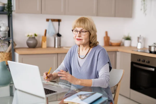 Современная зрелая женщина сидит перед ноутбуком за кухонным столом — стоковое фото
