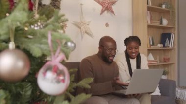 Oturma odasında Noel ağacıyla oturan ve dizüstü bilgisayara bakarken sohbet eden Afro-Amerikan kadın ve erkek pozu.