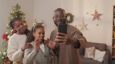 Genç Afro-Amerikan baba, anne ve 10 yaşındaki kızının, oturma odasında Noel ağacıyla cep telefonuyla konuştuğu el kamerası görüntüleri.