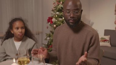 Üç çocuklu Afro-Amerikan ailenin Noel ağacıyla oturma odasında oturup Noel yemeğinden önce şükran duası ettiği bir sahne.