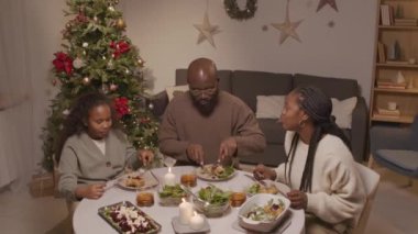 Afro-Amerikalı erkek, kadın ve kızın şenlikli oturma odasındaki yemek masasında oturup Noel yemeğinin tadını çıkarırken çekilmiş el kamerası görüntüleri.