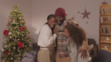 Orta boy mutlu bir Afro-Amerikan babanın heyecanlı karısı ve kızıyla sohbet ettiği ve Noel 'de eve döndükten sonra onları kucakladığı bir fotoğraf. Arka planda ışıklı Noel ağacı