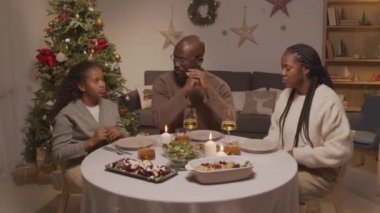 Afro-Amerikalı bir adam, bir kadın ve 10 yaşındaki bir kızın oturma odasındaki yemek masasında oturmuş Noel ağacını aydınlatıp şükran duasını okurken çekilen görüntüler.
