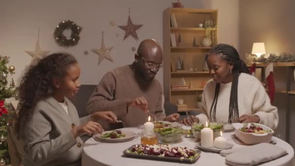 アフリカ系アメリカ人の少女 男と女の追跡ショット光と装飾されたクリスマスツリーと居心地の良いリビングルームでクリスマスディナーを食べる — ストック動画