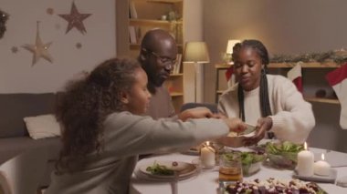Üç çocuklu Afro-Amerikan ailenin yemek masasında oturup Noel yemeklerini yerken rahat bir oturma odasında kutlama çekimleri.