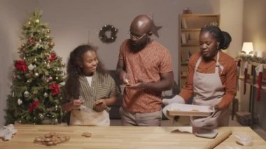 Mutlu Afro-Amerikan baba ve kızının, oturma odasında Noel ağacıyla zencefilli kurabiye süslerken, neşeli anne önlüklü olarak, taze pişmiş masayı...