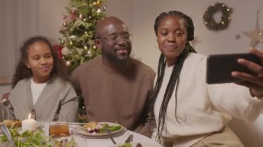 Orta boy mutlu bir Afro-Amerikan kızı, yemek masasında oturan ve Noel 'i birlikte kutlarken cep telefonuyla selfie çeken kadın ve erkek.