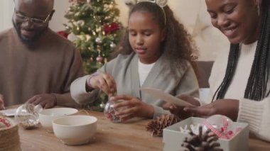 Orta boy mutlu Afrika kökenli Amerikalı baba, anne ve kız oturma odasında Noel ağacıyla oturup Noel hediyesi hazırlıyorlar. Cam takılar yapıp, kağıt imzalıyorlar.
