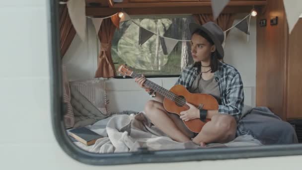 Tracking shot through window of camper young hipster woman in hat and checkered shirt sitting on cozy bed and playing music on ukulele