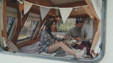 El kamerasıyla karavanın camından çekilen yavaş çekim mutlu bir kadın yatakta oturuyor ve yakışıklı bir kurt gibi okşuyor bir adam ukulele ile şarkı söylerken