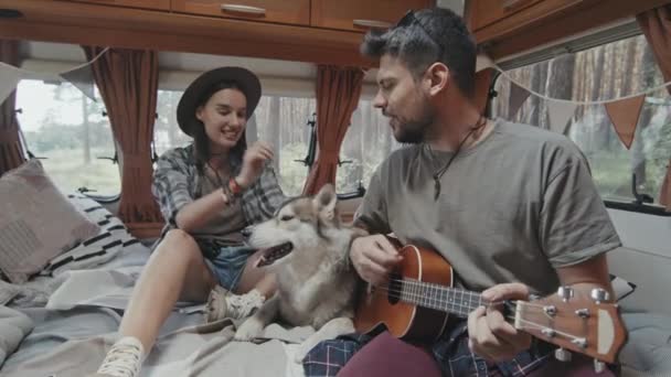 在舒适的露营车里 年轻人坐在床上 在Ukulele上唱歌 他快乐的女朋友坐在他身边边唱歌边爱抚可爱的哈士奇 — 图库视频影像