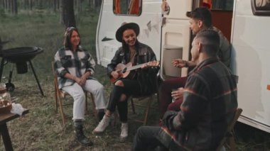 Kamp alanında arkadaşları için ukulele çalan genç bir kadının orta ölçekli çekimi. Beyaz karavan arkasına park etmiş.