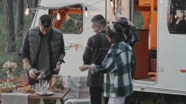 Kamp alanında piknik yapan mutlu çiftlerin yavaş takibi. Gülüyorlar ve masada oturuyorlar. Ormanda beyaz karavanın önünde duruyorlar.