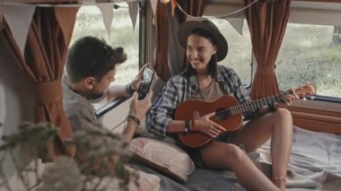 Erkek arkadaşı açık pencereden bakıp cep telefonuyla fotoğraf çekerken, karavanda oturup ukulele çalan mutlu genç bir kadının orta ölçekli görüntüsü.