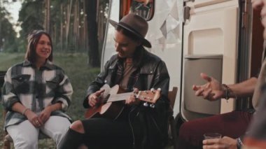 Karavanın önünde oturan ve neşeli arkadaşları şarkı söyleyip müzik dinlerken ukulele çalan genç kadının el kamerasındaki yavaşlığı.