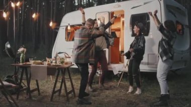 Yaz akşamları kamp alanında dans eden genç arkadaşların yavaş çekimleri. Beyaz karavan, rahat lambalar ve yemek dolu piknik masası var.
