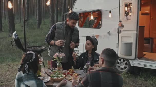 幸せな若い男性と女性の遅いテーブルに座って キャンプ場でピクニックをしています 乾杯と言ってアルコールのガラスを持つ男は その後 彼の友人とグラスをクリックします 横に停めた白いキャンパー — ストック動画