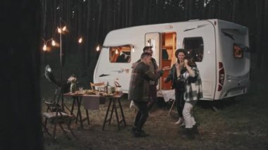 Yaz akşamları rahat kamp alanında dans eden mutlu gençlerin yavaş çekimleri. Arka planda park etmiş beyaz bir karavan.