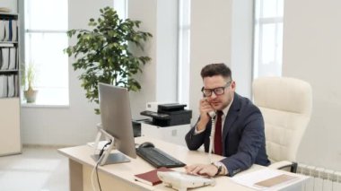 Erkek yöneticinin ofis masasında oturup telefon görüşmesi yaptığı orta boy bir fotoğraf. Kadın kişisel asistan odaya giriyor ve talimat verirken not alıyor.