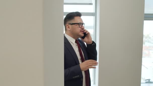 オフィスの窓のそばに立って携帯電話で話しているスーツと眼鏡で厄介な男性Ceoの追跡ショット — ストック動画