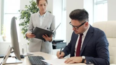 Takım elbiseli ve gözlüklü ciddi bir erkek yöneticinin ofis masasında oturup evrakları imzalarken, sonra da onları kadın kişisel asistana verirken orta boy resmi.