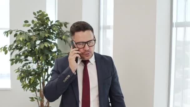 手持中镜头的沮丧商人身穿西服 戴着眼镜 在办公室用手机进行了热烈的交谈 然后怒气冲冲地挂断电话 — 图库视频影像