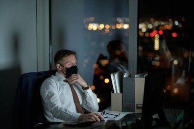 Koruyucu maskeli genç beyaz yakalı işçi bilgisayar ekranına bakıyor.