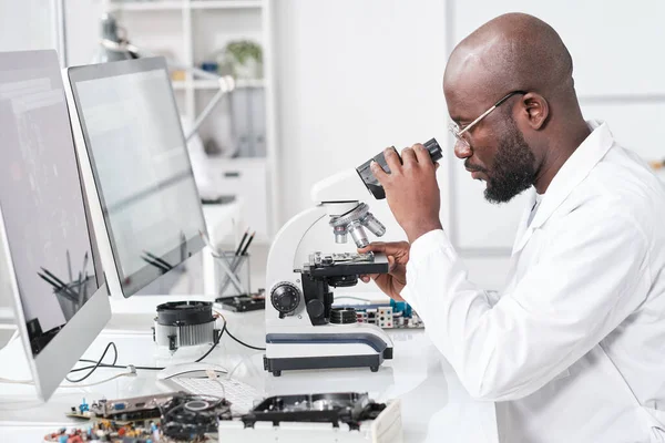 Oldalnézet fiatal afrikai tudós fehérköpenyben tanulmányozza mikrochip jellemzői — Stock Fotó