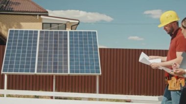 Kadın yöneticinin dosya ve erkek mühendis ile PAN görüntüsü. Belge ile konuşuyor ve banliyö bölgesinde kurulmuş güneş panellerinin önünden geçiyor.
