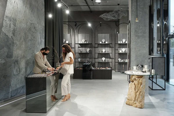 Jovem comprador feminino na máscara escolhendo jóias enquanto o assistente de loja masculino ajudá-la — Fotografia de Stock