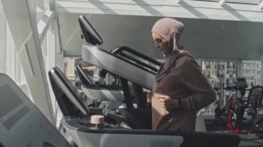 Koşu bandında koşan tesettürlü genç Müslüman kadın spor yaptıktan sonra dur düğmesine basıp su içiyor.