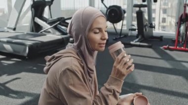 Başörtülü güzel Müslüman kadın spor salonunda yerde oturuyor ve sallanan şişeden su içiyor.