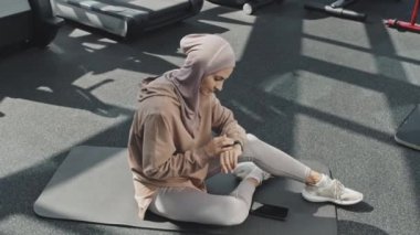 Spor salonundaki tesettürlü Müslüman kadının spor salonundaki mindere oturduğu ve spor programı uygulamasını akıllı sete ayarladığı görüntüyü yakınlaştır