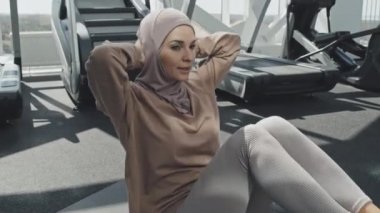 Başörtülü genç Müslüman kadın spor salonunda kas egzersizi yaparken minder üzerinde Ziyaretçi-oturma egzersizi yapıyor.