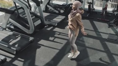 Yüksek açılı çekimde tesettürlü ve spor kıyafetli bir kadın spor salonunda egzersiz yaparken ipten atlarken görülüyor.